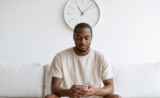 man looking at his phone with a clock in the wall to represent the time the dermatologist takes to respond via miiskin